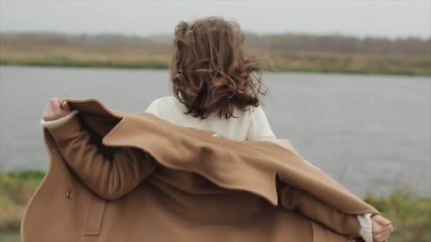 Eine hübsche junge Frau mit Brille steht auf dem Hintergrund des Flusses und zieht ihren Mantel von den Schultern und lächelt in die Kamera. Zeitlupe — Stockvideo