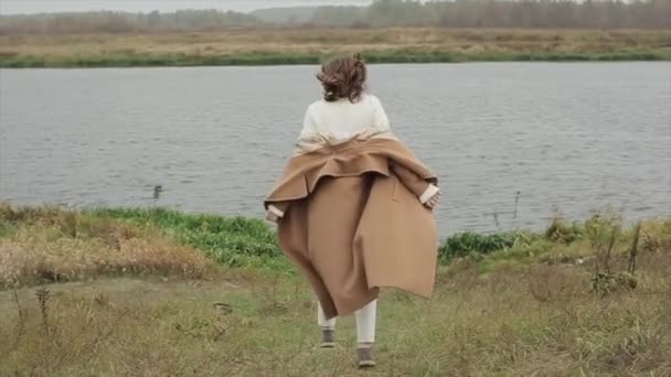 Приваблива молода дівчина в коричневому пальто відправляється на річку в осінній вітряний день і обертається навколо і дивиться на камеру. Повільний рух — стокове відео