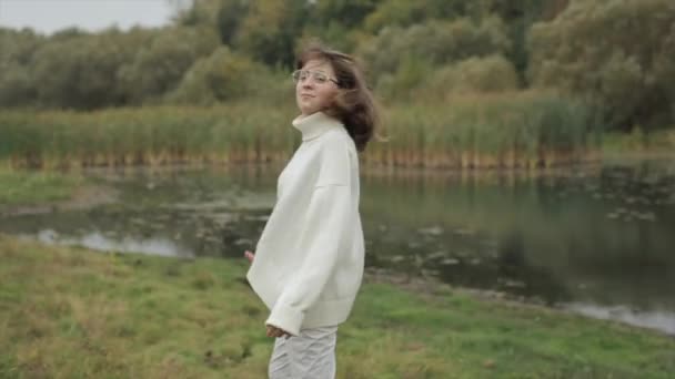 Мила молода дівчина в білому одязі йде до річки в осінній день і обертається і дивиться на камеру посміхаючись — стокове відео