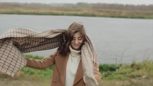 Güzel bir genç kadın nehrin arka planında duruyor ve boynuna bir eşarp takıyor ve kendisini bir paltoya sarıp gözlerini kapatıyor. Yavaş çekim — Stok video