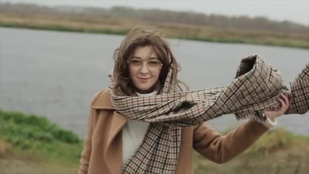 Una jovencita sonriente con un abrigo se levanta sobre el fondo del río y endereza su cabello, desaliñado del viento. Movimiento lento — Vídeos de Stock