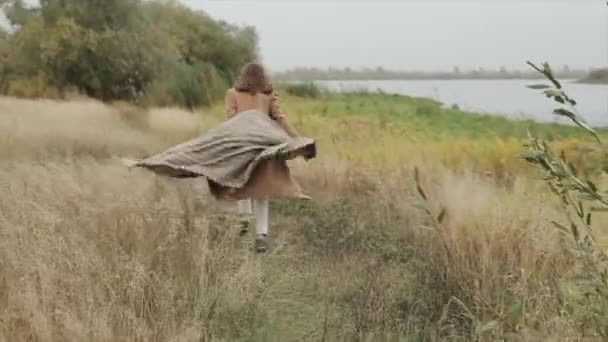 An attractive young girl goes along the river bank in an autumn day and holds in her outstretched hands a large scarf fluttering in the wind. Slow motion — Stock Video