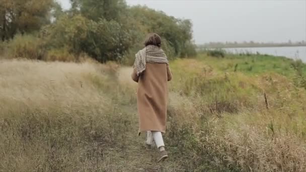 Чуттєва молода жінка в окулярах стоїть з закритими очима на березі річки в вітряний осінній день і тримає в роті лезо трави. Повільний рух — стокове відео