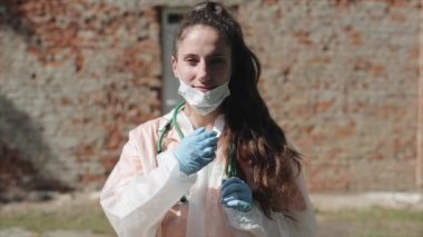 Bir yaz günü sokakta durup kameraya bakan koruyucu beyaz tulumlu ve sağlık maskeli bir kadın doktor portresi. Koronavirüs salgını. Yavaş çekim