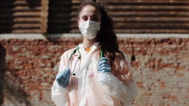 Een jonge verpleegster in beschermende uitrusting tijdens de COVID-19 staat op straat en houdt met haar handen een fonendoscoop om haar nek. Middelmatig schot. Langzame beweging — Stockvideo