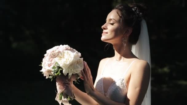 Una sposa delicata in un velo e in un abito aperto lucido sta con gli occhi chiusi al sole di profilo e tocca il bouquet. — Video Stock