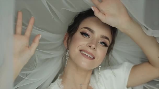 Una joven novia sensual con hermoso maquillaje está rodeada por un velo blanco transparente y pone su dedo en sus labios. Primer plano — Vídeos de Stock