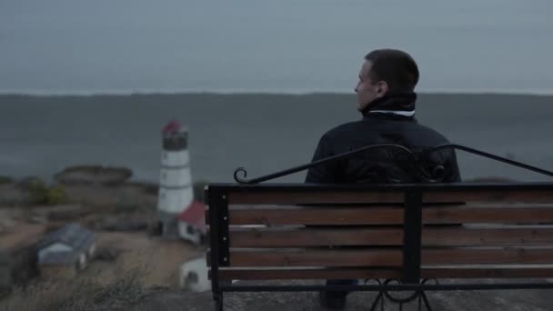 En ung man i svart jacka sitter på en bänk och beundrar havet och fyren vid foten av kullen — Stockvideo
