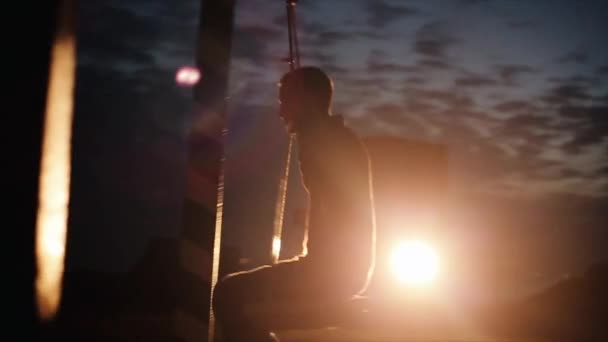 Ein junger Mann fährt am späten Abend im Scheinwerferlicht eines Autos auf einer Schaukel und lächelt, als erinnere er sich an seine Kindheit. Zeitlupe — Stockvideo
