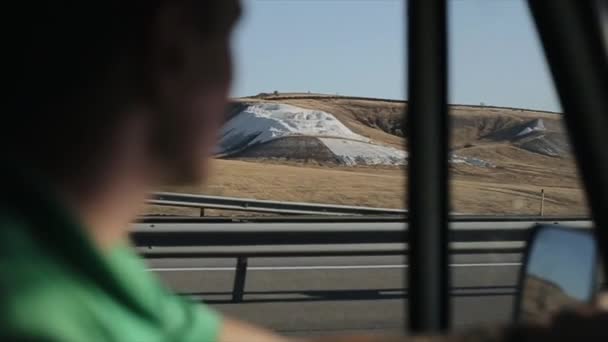 Молодой человек в зеленой футболке едет на своем фургоне по дороге и восхищается белыми холмами за окном. Внутри машины. — стоковое видео