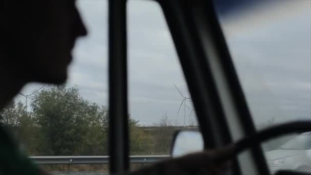 Молодой человек едет на своем фургоне по шоссе, и ветряные турбины медленно вращаются за окном. Размытый передний план — стоковое видео