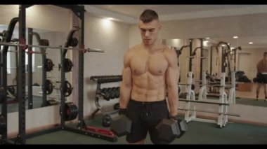 Genç ve güçlü bir adam spor salonunda antrenman yapıyor ve omuzları ve trapezius kası için dambıl omuz silkiyor.