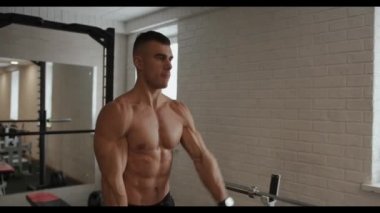 Genç kaslı adam omuz kaslarını çalıştırıyor ve kollarını spor salonunda ağır dambıllarla sırayla öne doğru kaldırıyor.