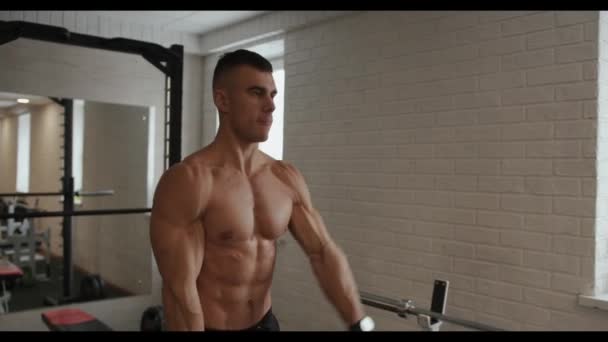 Jeune homme musclé travaille les muscles de l'épaule et lève les bras alternativement en face de lui avec de lourds haltères dans la salle de gym — Video