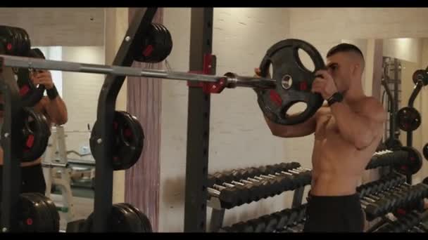 Jonge gespierde atleet in zwarte sportbroek op een training in de sportschool en hangt pannenkoeken op een lange halter voor krachtoefeningen — Stockvideo