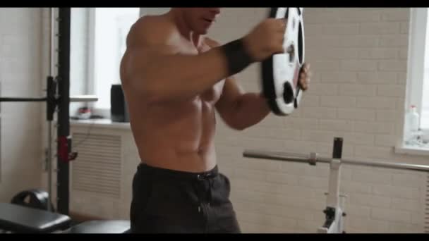 En ung bodybuilder tog i gymnastiksalen og roterer pandekager vægtstænger omkring hovedet, pumpe op pressen og skulderbæltet – Stock-video