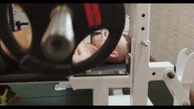 Spor salonundaki bir bankta uzanırken ağırlık kaldırma halteri yapan güçlü bir genç adam trisepsi ve omuz kaslarını pompalıyor.