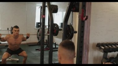 Genç kaslı adam spor salonunda antrenman yapıyor ve sırtında halterle bacağının ön kısmını pompalıyor.