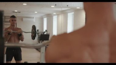 Çıplak gövdeli kaslı genç adam spor salonunda aynanın önünde duruyor ve halterle pazılar için egzersiz yapıyor.