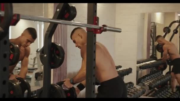 Jonge gespierde man met een naakte romp op een training in de sportschool en kiest een pannenkoek voor een lange halter hangend aan een rek — Stockvideo