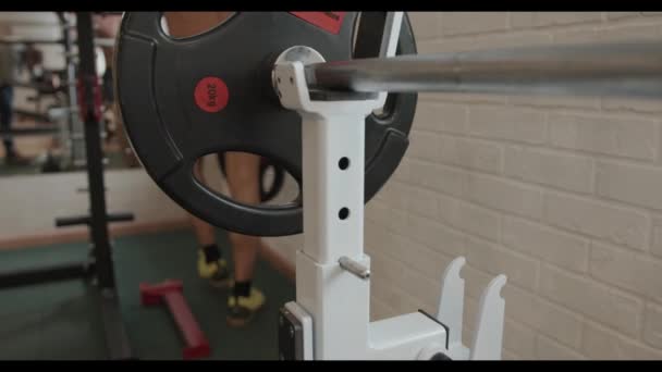 Joven fisicoculturista con un torso desnudo entrenando en el gimnasio y quita los panqueques de una barra que está en un bastidor — Vídeo de stock