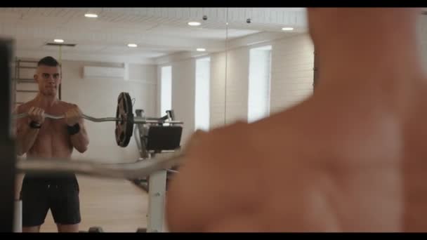 Młody, umięśniony mężczyzna z nagim tułowiem stoi przed lustrem w siłowni i wykonuje ćwiczenia na bicepsy ze sztangą — Wideo stockowe