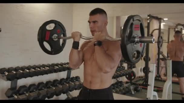 Jovem fisiculturista em calções esportivos pretos bombeia bíceps com um z- barbells enquanto está em frente a um espelho no ginásio — Vídeo de Stock