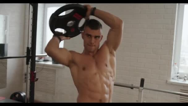 Młody muskularny mężczyzna podczas treningu na siłowni ćwiczy na pasku naramiennym i obraca sztangę na naleśnikach wokół głowy — Wideo stockowe