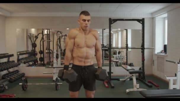 Een jonge man met een naakte romp training in de sportschool en doet een oefening op de trapezius spier tilt zijn schouders met halters in zijn handen — Stockvideo