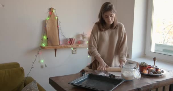 Linda chica joven lanza masa de jengibre para las vacaciones de Navidad en una mesa de madera en una cocina decorada y acaricia a su perro — Vídeo de stock
