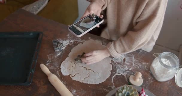 Ένα νεαρό κορίτσι κόβει μελόψωμο από ζύμη με ένα σιδερένιο καλούπι σε σχήμα μικρού ανθρώπου για τα Χριστούγεννα και απογειώνει τη διαδικασία σε ένα κινητό τηλέφωνο. Άποψη από ψηλά — Αρχείο Βίντεο