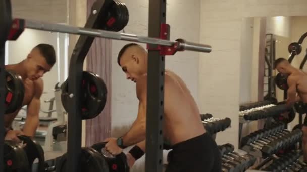 Junge Bodybuilderin mit nacktem Oberkörper lehnt an Metallhanteln und macht Handstand vor einem Spiegel im Fitnessstudio — Stockvideo
