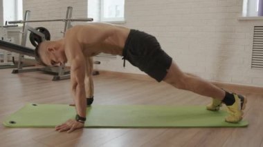 Spor salonunda ve paspasın üzerinde vücut geliştirme eğitimi alırken dirseklerini dizlerinin üstüne çekip kalçalarını kaldırır.