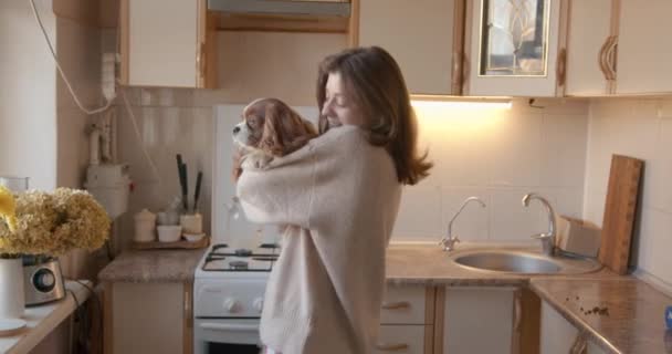 Ένα χαριτωμένο γούνινο σκυλί κάθεται στην αγκαλιά μιας νεαρής γυναίκας και ακουμπά το κεφάλι του στον ώμο της ιδιοκτήτριας που χορεύουν μαζί στην κουζίνα. Αργή κίνηση — Αρχείο Βίντεο