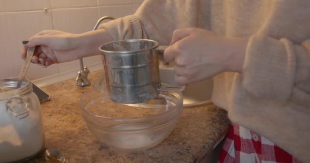 En ung flicka i röda och vita byxor matlagning pepparkakor för julhelgen och sållar mjöl i en glasskål — Stockvideo