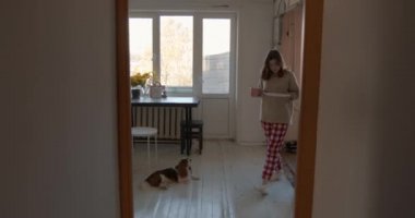 Ev kıyafetleri içindeki sevimli genç bir kız elinde büyük bir tabak ve bir kupayla mutfaktan çıkıyor ve köpeği peşinden koşuyor.