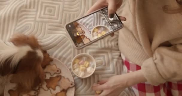 Una giovane ragazza con il suo cane siede su un letto con un piatto di pan di zenzero e una tazza di cacao con marshmallow e fotografa il cibo su un telefono cellulare. Vista dall'alto — Video Stock