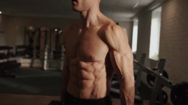 Genç kaslı adam spor salonunda antrenman yapıyor ve pazılarını sallıyor. Kollarını ağır halterlerle esnetiyor.