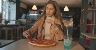 Uzun sarı saçlı çekici bir kız bir kafede bir masada oturuyor ve yüzünün bir kısmını bir dilim pizzayla kaplıyor.