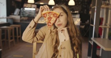 Genç bir sarışın kız bir kafede pizza yiyor ve gülümsemesiyle yüzünün bir kısmını kaplayan bir dilim pizzayı tutuyor.