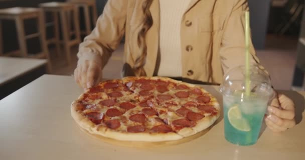 Een mooi jong meisje in een beige jas zit in een café en eet een pizza met haar handen en drinkt een blauwe cocktail door een rietje — Stockvideo