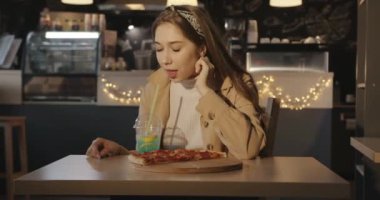Sarı uzun saçlı neşeli genç kız bir kafede pizza yer ve pipetle soğuk mavi bir kokteyl içer.