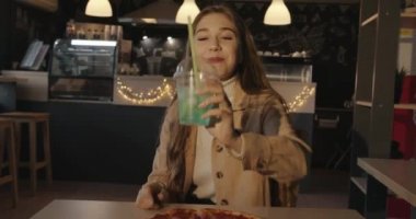 Çekici sarışın kız bir kafede elinde pizzayla oturuyor ve elinde kameraya buz ve limonlu mavi bir kokteyl tutuyor.