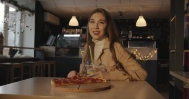 Uzun sarı saçlı, mavi kokteylli bir kafede pizza yiyen ve kameraya göz kırpan tatlı bir genç kız.