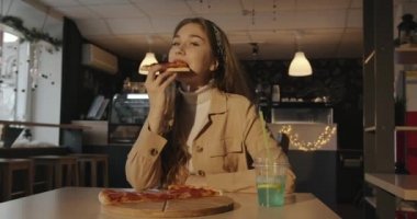 Beyaz kazak ve bej ceketli neşeli genç kız bir kafede pizza yer ve pipetle mavi bir kokteyl içer.