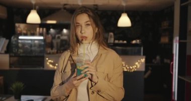 Uzun sarı saçlı güzel bir kız bir kafede dikiliyor ve limon ve buzlu mavi bir kokteyl içiyor.