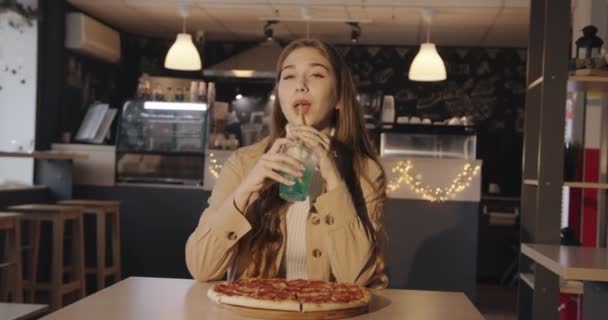 Una agradable jovencita con chaqueta beige come pizza en un café y bebe un cóctel azul con limón a través de una paja y guiño — Vídeos de Stock