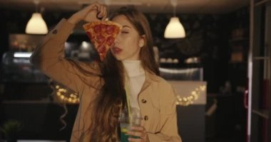 Uzun sarı saçlı çekici bir genç kız bir kafede bir bardak kokteyl ile duruyor ve yüzünün yarısını kaplayan bir dilim pizzayı elinde tutuyor.