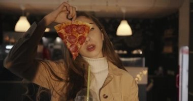 Şehvetli genç sarışın kız bir kafede elinde bir kokteyl ile duruyor ve yüzünün bir kısmını pepperoni pizzasıyla kapatıyor.