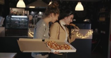 İki güzel, uzun saçlı kız bir kafede durup bir kutudan pizza yiyor ve gülüyorlar. Yavaş çekim